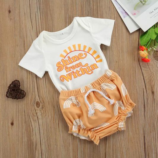A 2 piece set of a baby onesie and bloomers are shown. The onesie is white with orange words that say "Shine From Within" in a cute font with sun rays coming out of it. The bottoms are orange with white rainbows on them.