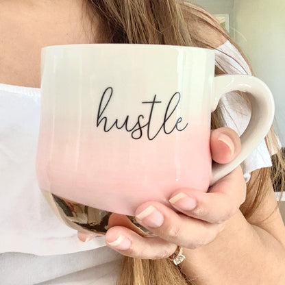 Woman holding light pink ombre mug with a gold bottom and gold handle that says Hustle